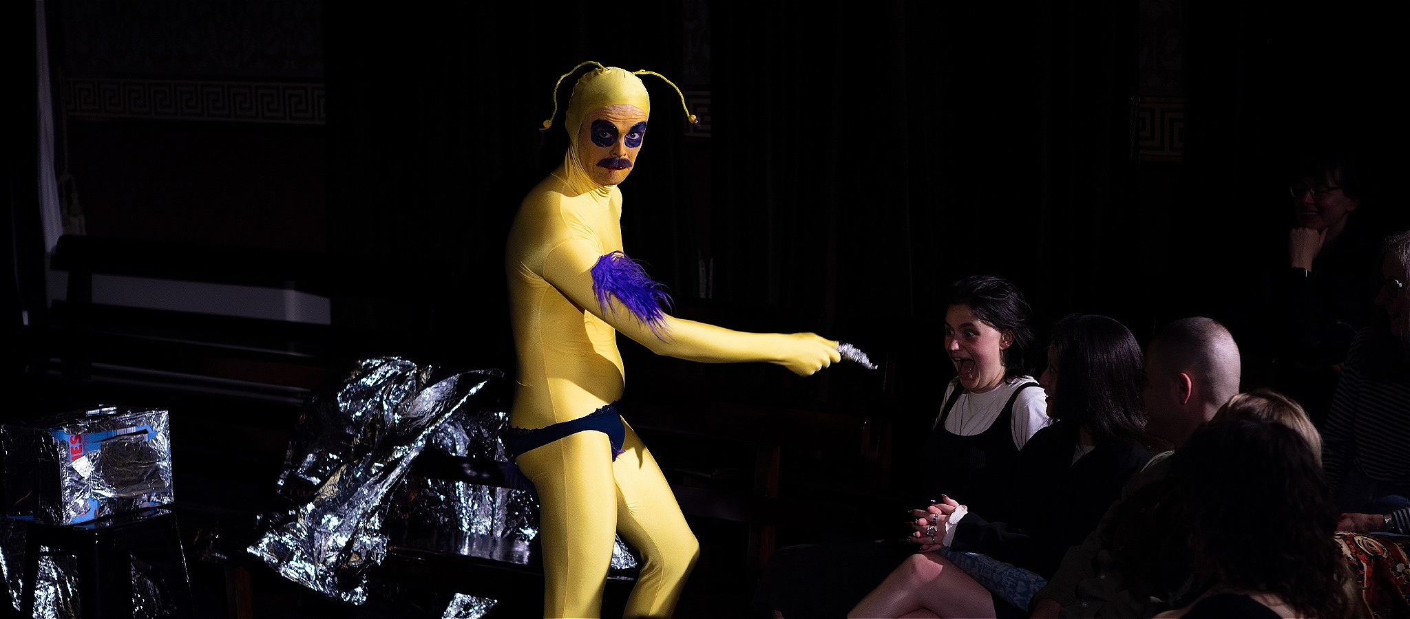 84BY, the yellow alien with purple fur, confronts a laughing audience member mid-performance.