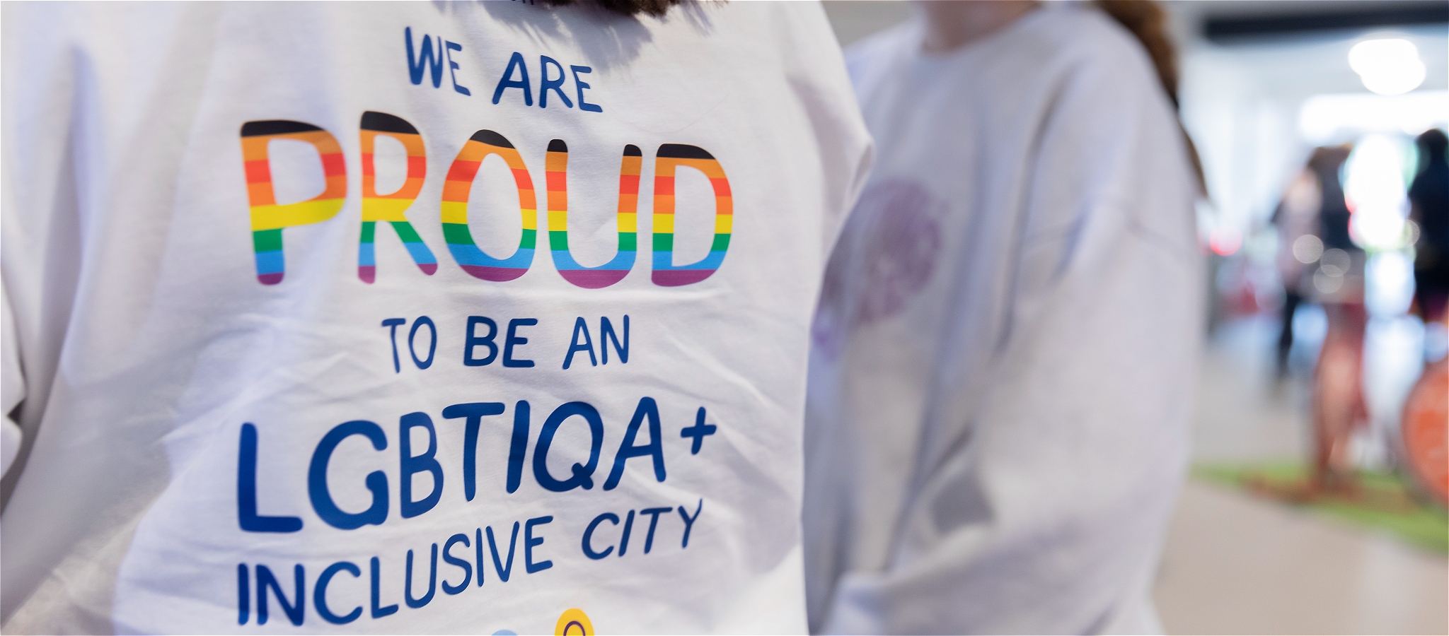 The back of a white t-shirt with rainbow-coloured text that says - We Are Proud To Be An LGBTIQA+ Inclusive City